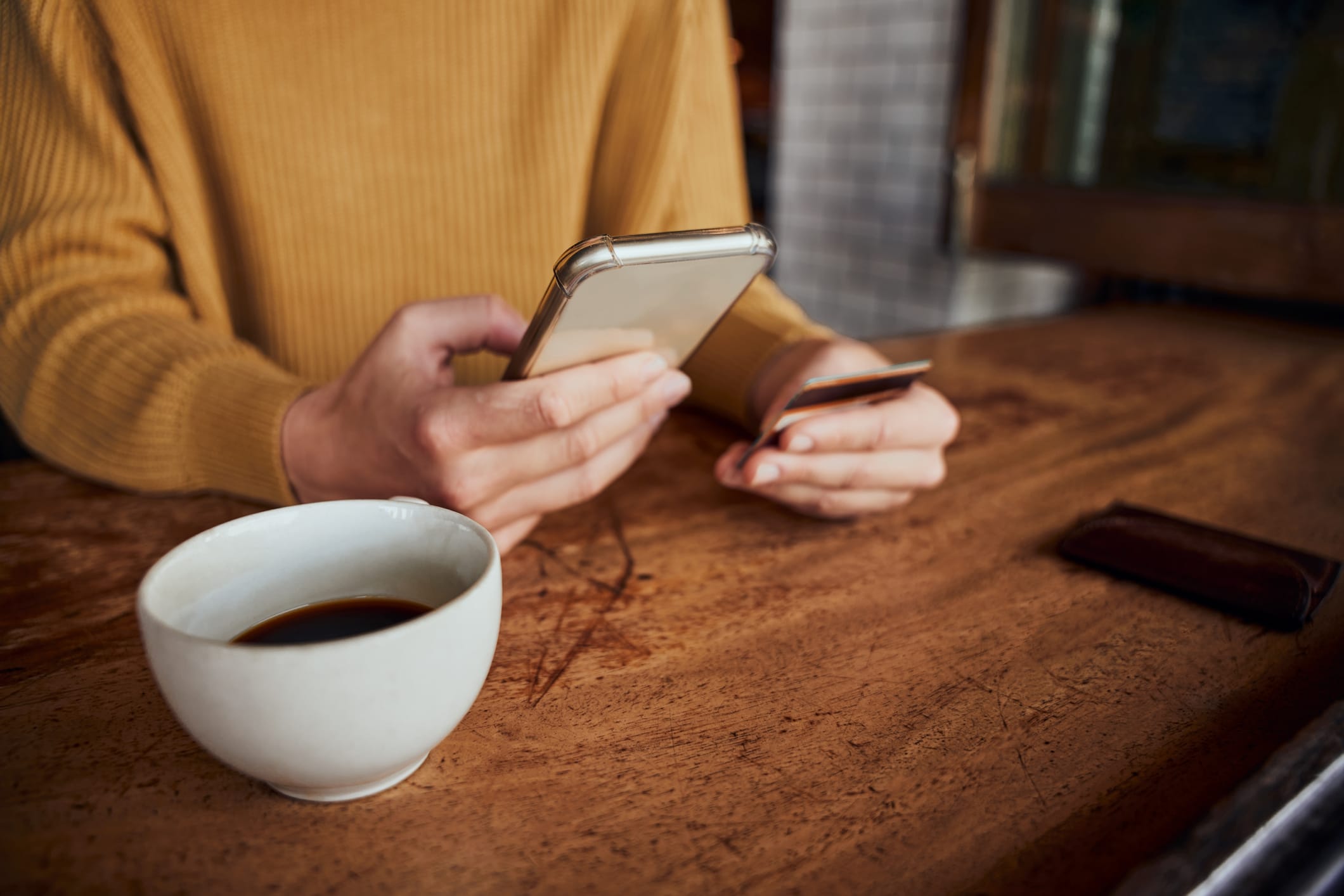 Closeup of young female hands holding smartphone and bank card while making online payment in cafe drinking coffee