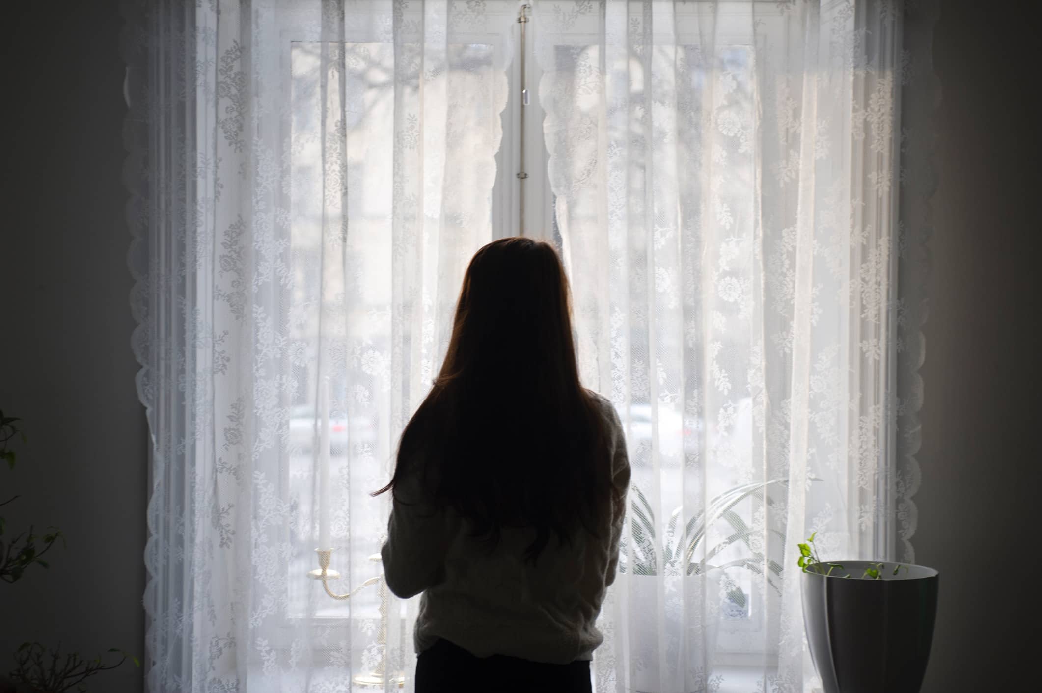 Rear View Of Woman Looking Through Window While Standing At Home
