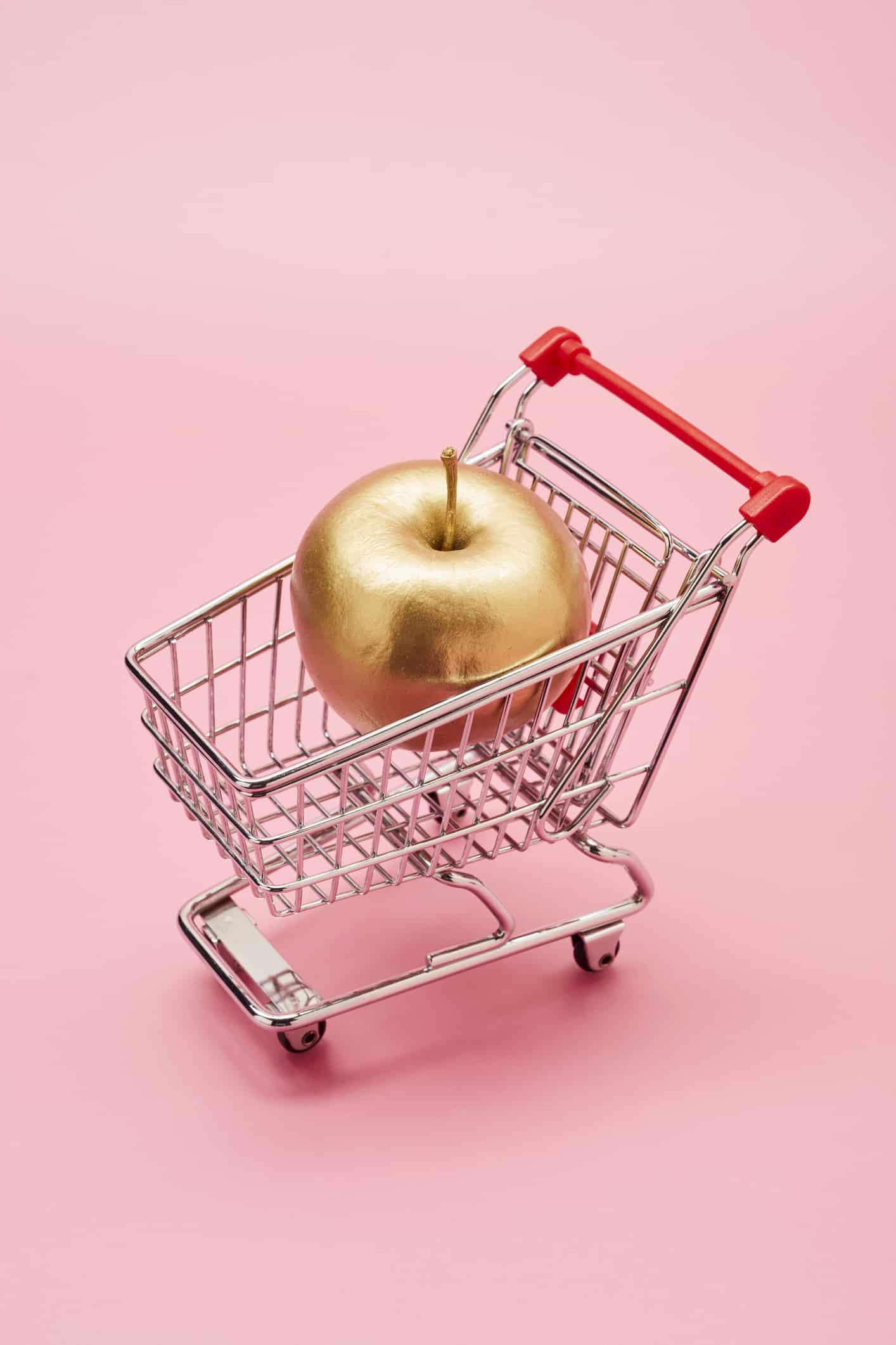 Still life of a small shopping cart and a golden apple on pink background