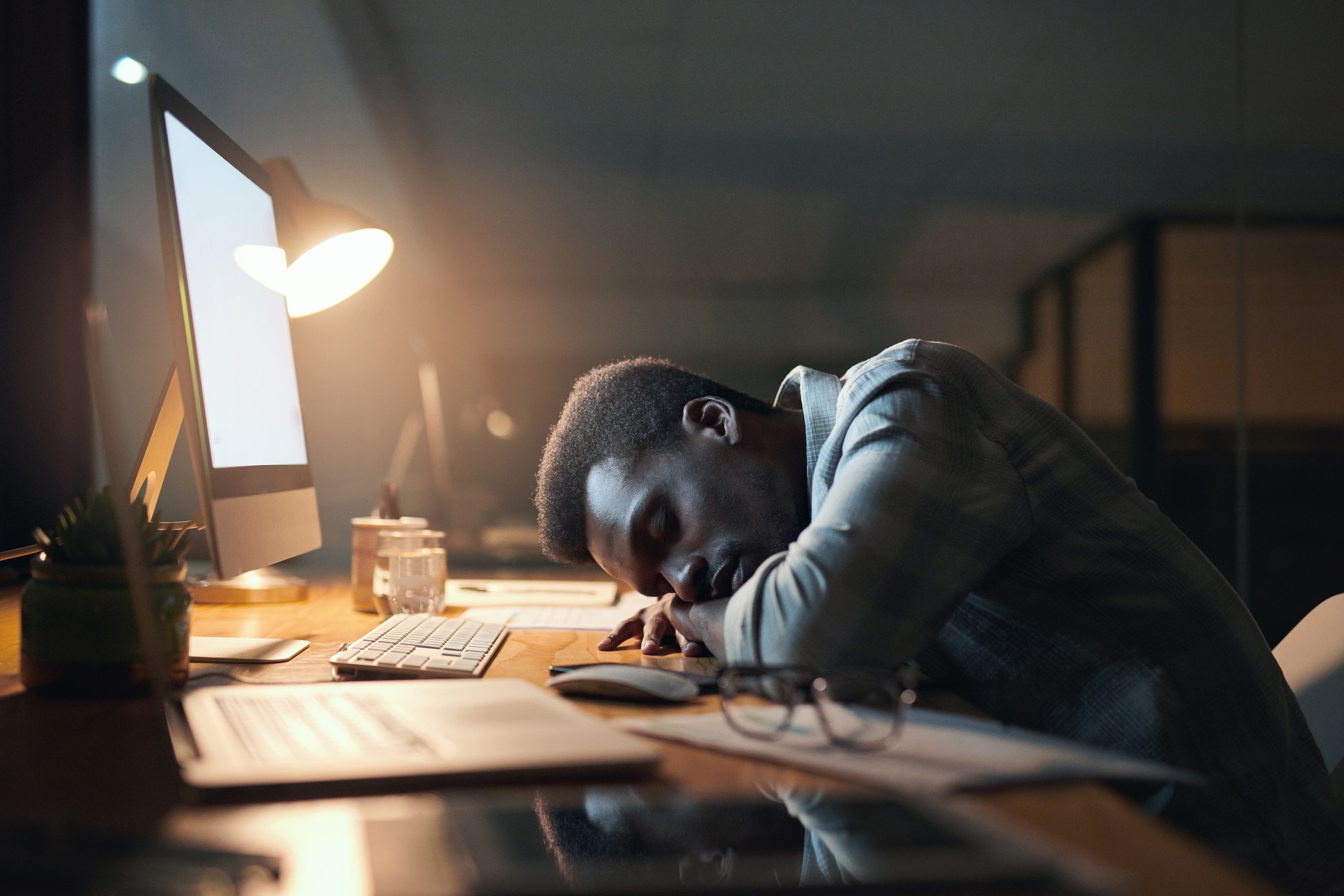 Tired Black man, sleeping and burnout from working at night by office desk suffering stress or overworked. Exhausted African American male asleep on table by computer for long work hours at workplace