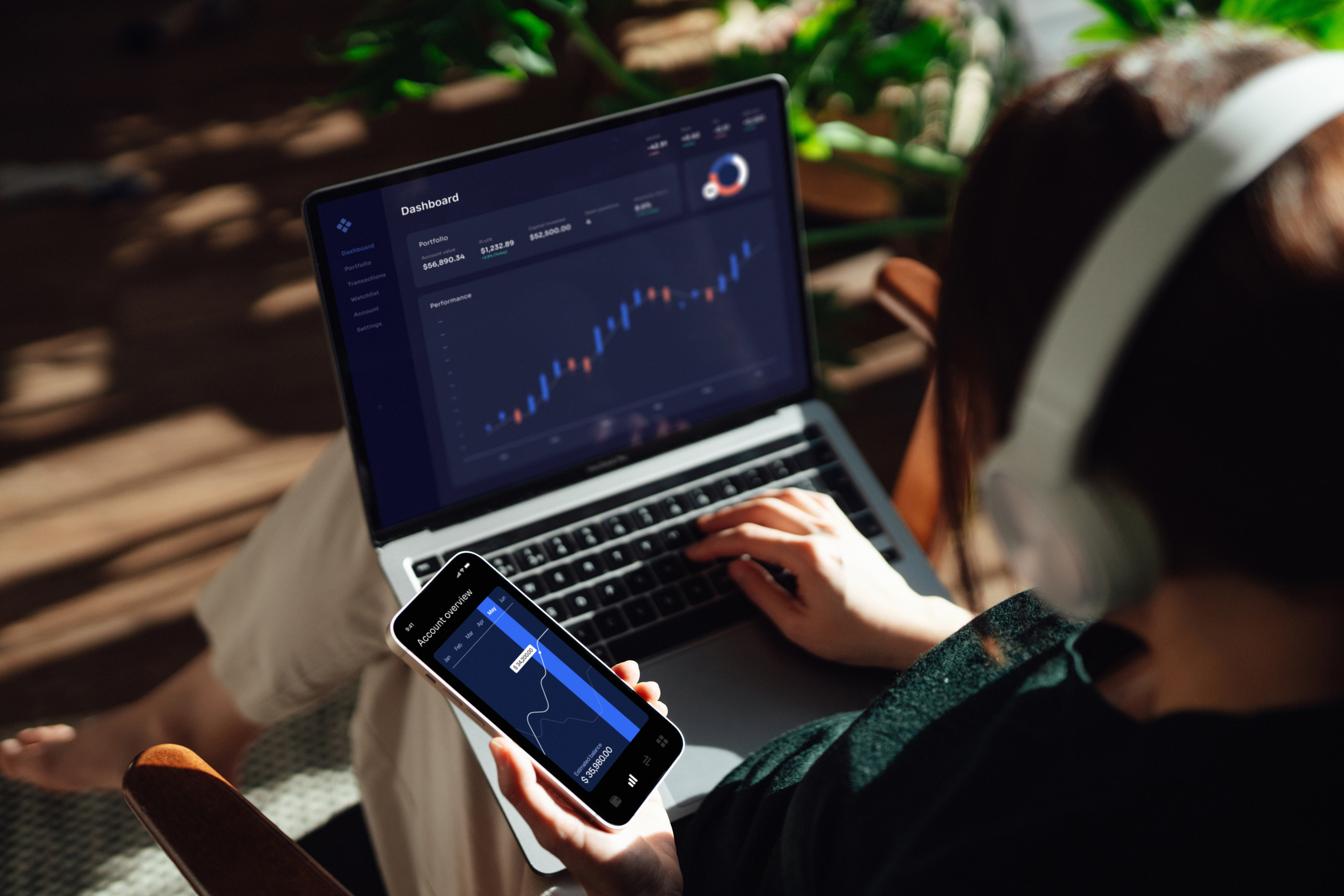 Over the shoulder view of young woman making financial plans and investment analysis via laptop and smartphone